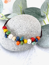 Load image into Gallery viewer, A beaded bracelet with colorful, textured round beads in red, yellow, green, blue, orange, white, and light blue, strung on a dark brown cord. The bracelet is displayed on a round gray stone with eucalyptus leaves in the background.
