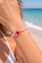 Load image into Gallery viewer, Close-up of a pink beaded bracelet with a heart charm on a person's wrist at the beach.
